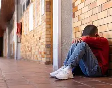 Child sitting alone on the floor of a school hallway with head down and arms over their knees.